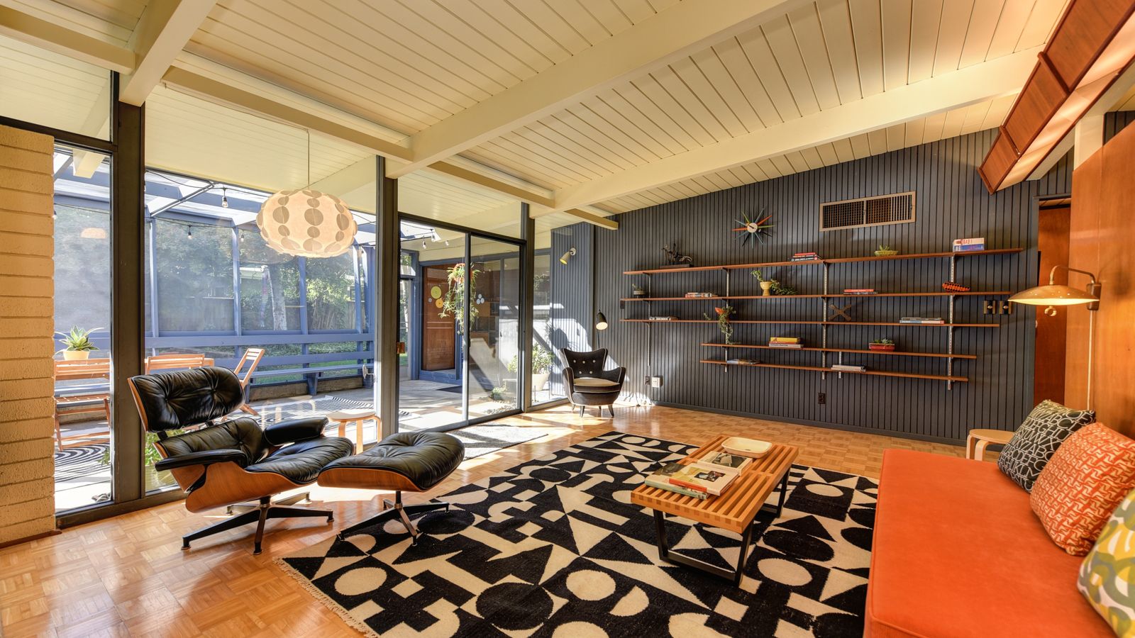 Mid-century post-and-beam living room with a floor-to-ceiling glass wall, exposed wood beams, and an Eames lounge chair lit by a single floor lamp